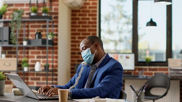 Male consultant with face mask answering landline phone call, talking to manager about business statistics. Using office telephone to have remote conversation on telework dial line. Tripod shot.