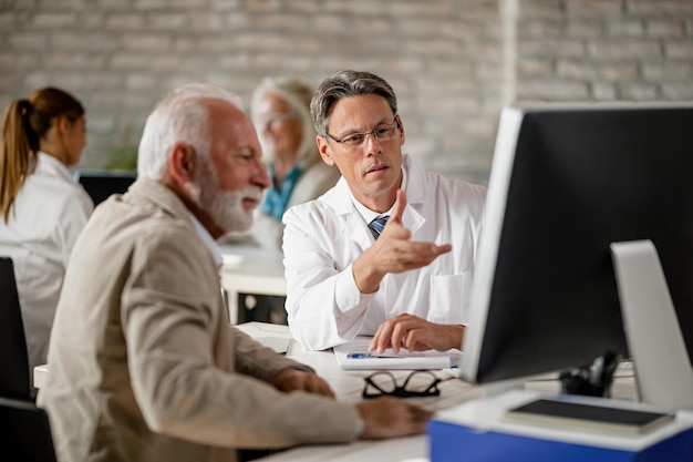 Male doctor explaining to senior patient medical insurance policy while using computer during consultations at clinic