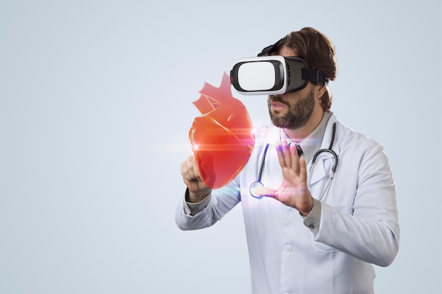 Male doctor on a gray background using a Virtual Reality Glasses, looking at a virtual heart.