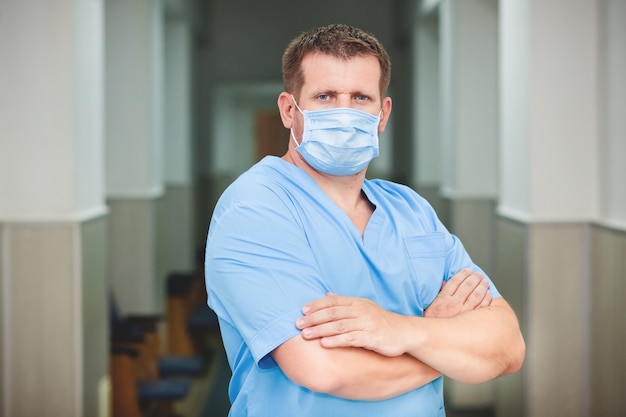 A male doctor in a medical mask on his face and uniform stands in the hallway of the clinic