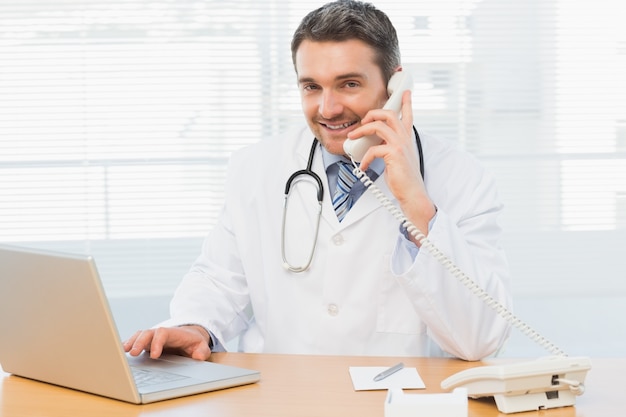 Male doctor using laptop and phone in medical office