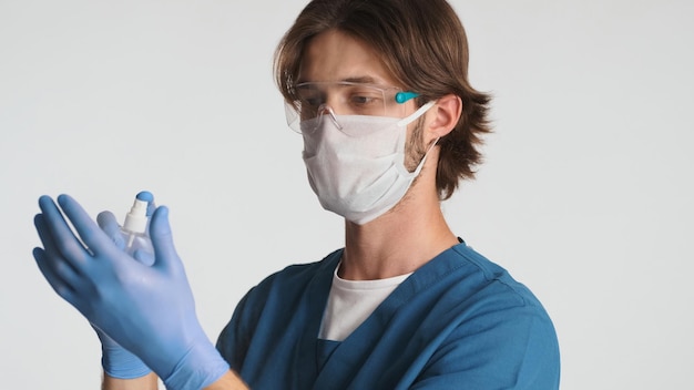 Male doctor wearing medical mask and gloves using antiseptic spray preparing for a working day in hospital Young intern in uniform and protected glasses standing