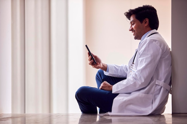 Male Doctor Wearing White Coat Sitting On Floor In Hospital Corridor Text Messaging On Mobile Phone