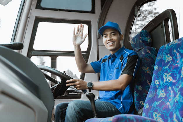 A male driver in uniform smiles at the camera as he sits down and waves at the bus