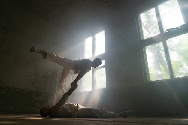 Male duo making acrobatic tricks wearing costume of insane people in abandoned room with sun rays from windows.