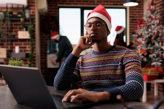 Male employee with santa hat using laptop in festive decorated workplace. Working on project in business office with christmas tree and decorations, seasonal holiday ornaments.