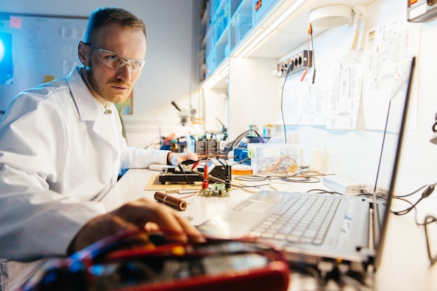 Male engineer in white robe using laptop for work in electronic laboratory