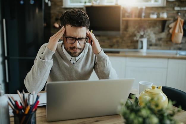 Male entrepreneur holding his head in pain while working on laptop at home