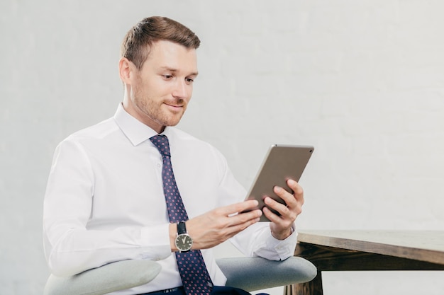 Male executive manager in white shirt and tie holds tablet computer searches necessary information in internet connected to wifi in office People business and modern technologies concept