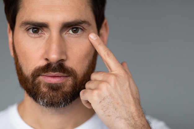 Male facial skincare Handsome caucasian bearded man touching face caring for skin under eyes grey background