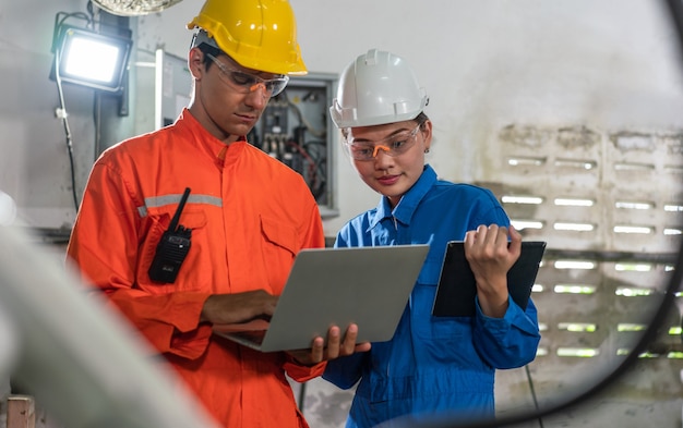 Male and female automation engineers wear a uniform with helmet safety inspection control a robot arm welding machine with a laptop in an industrial factory. Artificial intelligence concept.