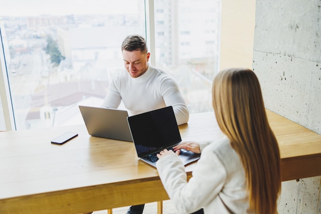 Male and female colleagues are working at a wooden table on laptops and talking Concept of business cooperation and teamwork Young colleagues in the office Modern successful people