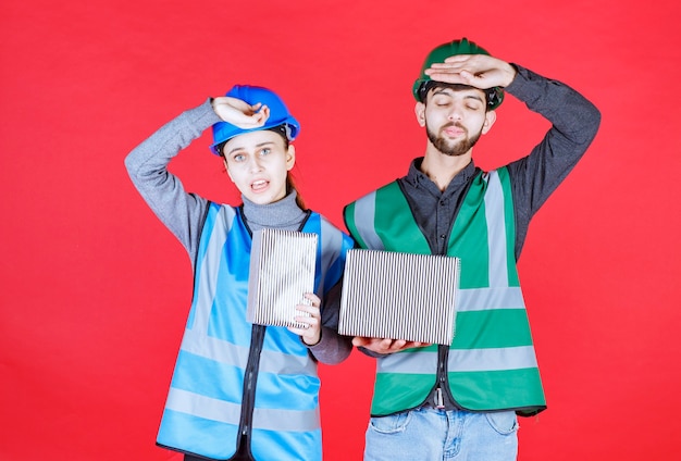 Male and female engineers with helmets holding silver gift boxes and look tired.