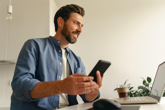 Male freelancer holding phone while working on laptop on home kitchen distance work concept