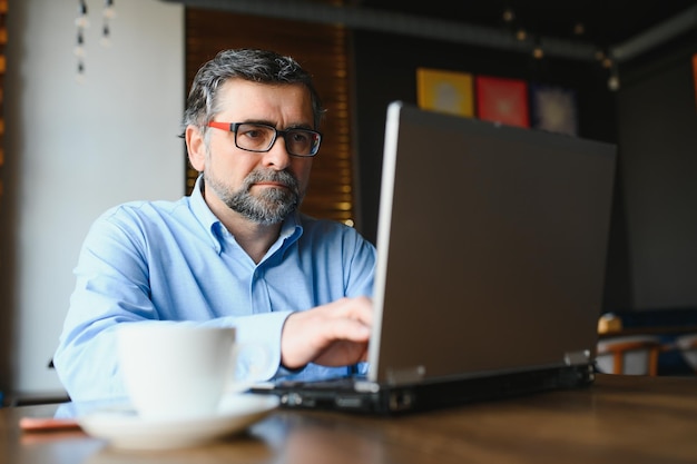 Male freelancer is working in a cafe on a new business project Sits at a large window at the table Looks at a laptop screen with a cup of coffee