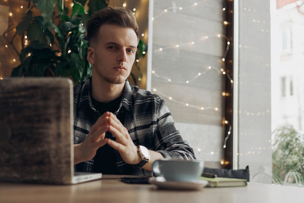 male freelancer looking at camera while sitting at table with laptop during work in cafe