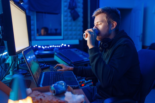 Male gamer drinking an energy drink at his workplace with laptop and desktop PC, gaming night lifestyle. Computer games player in his room with neon light, streamer