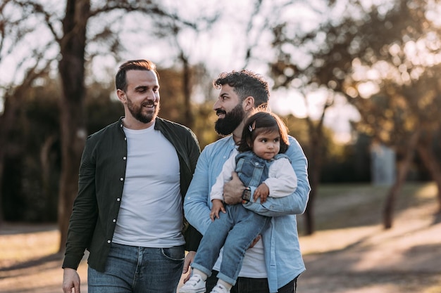 Male gay couple walk in the park in love with their daughter in their arms