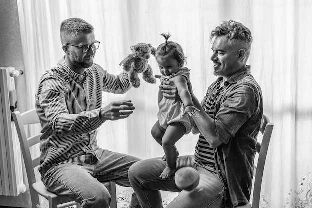 Male gay couple with adopted baby girl at home Two handsome fathers playing with their daughter