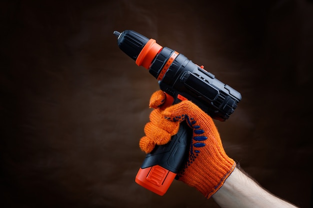 Male gloved hand holding an electric screwdriver on a dark brown background.