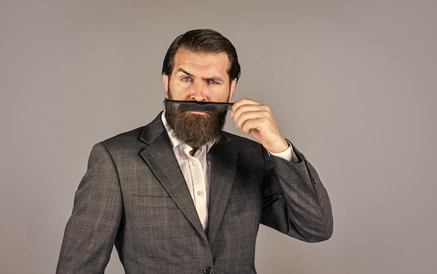 Male grooming concept mature businessman in suit brutal hipster with moustache in barbershop barber in hairdresser salon hair tool fashion and beauty bearded man brush beard Personal care