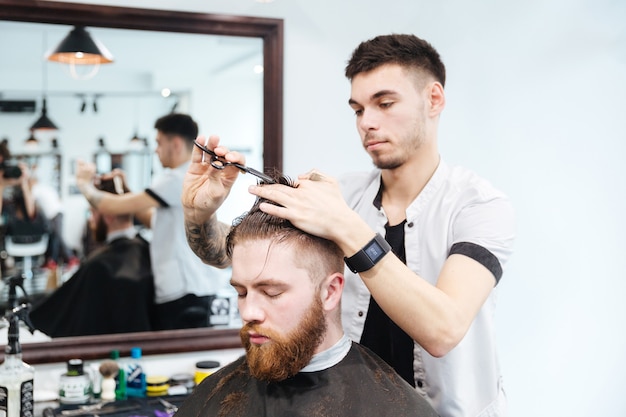 Male hairdresser cutting hair with scissors to his client in barbershop