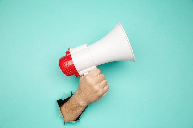 Male hand arm hold megaphone isolated through torn blue background.