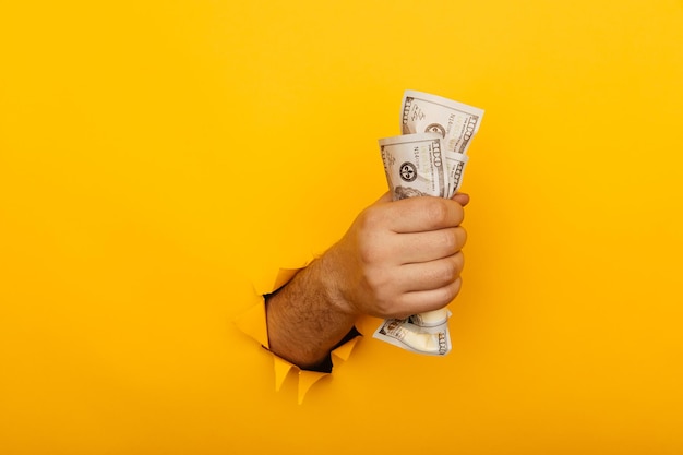 Male hand holding crumpled dollar banknotes through hole in yellow background