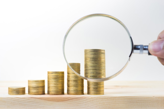 Male hand holding magnifying glass with coin stacked on wood table