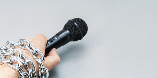 Male hand holding microphone tied with chains on grey background with copy space Banner with place for text