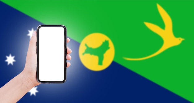 Male hand holding smartphone with blank on screen on background of blurred flag of Christmas Island Closeup view