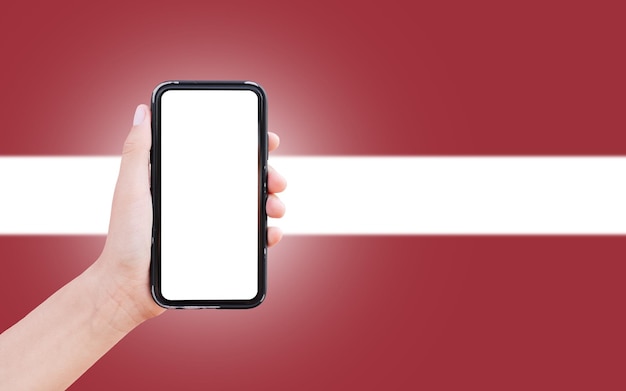 Male hand holding smartphone with blank on screen on background of blurred flag of Latvia Closeup view