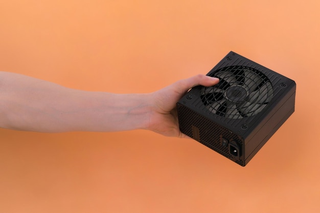 The male hand holds a cooler of computer power supply on an orange background Unit Computer Plug Black Box Equipment Hardware Source Device Part Metal Grey PC Powerful