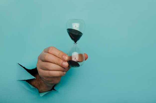 Male hand holds hpurglass in torn hole of blue background