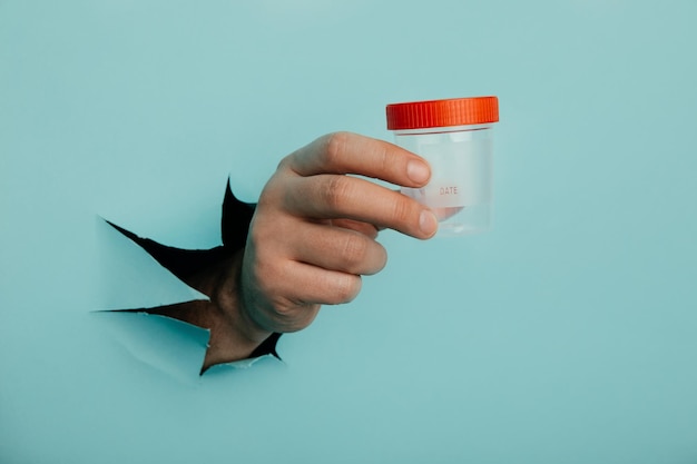 Male hand holds a plastic jar for urine tests the hand is sticking out of a torn hole in a blue background