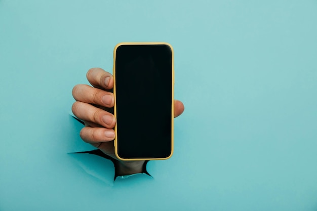 A male hand sticking out of a hole from a blue background holds a smartphone with a black screen