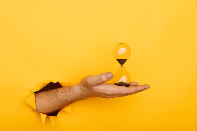 Male hand with hourglass through a hole of yellow background