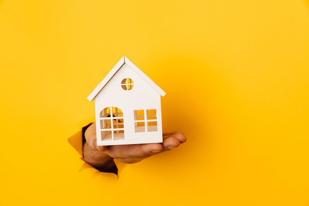 Male hand with mini house through a hole in yellow paper background