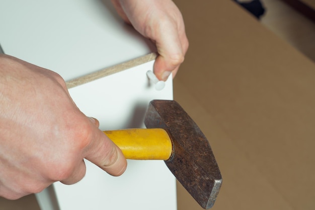 Male hands hold metal hammer with yellow plastic handle driving dowel assembling white drawer of dresser or wardrobe