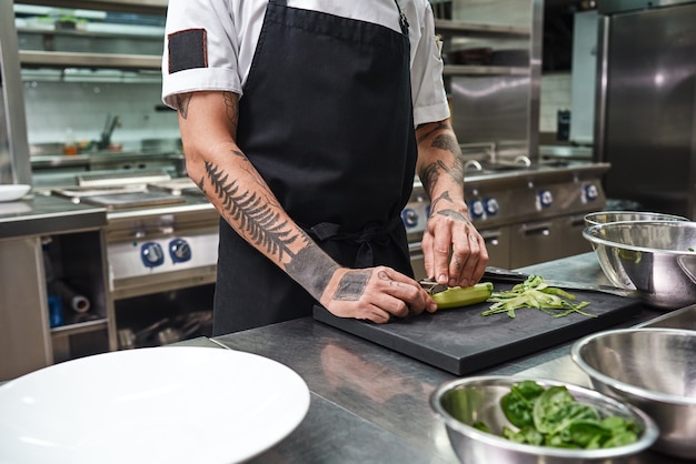 male hands with beautiful tattoos peeling cucumber for salad while standing in a restaurant kitchen