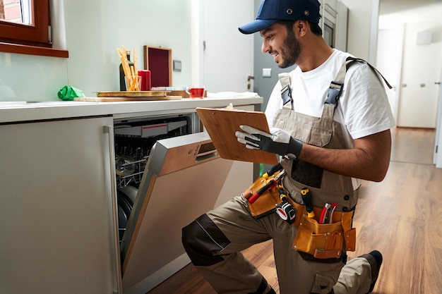 Male handyman sitting near dishwasher and writing on clipboard in kitchen