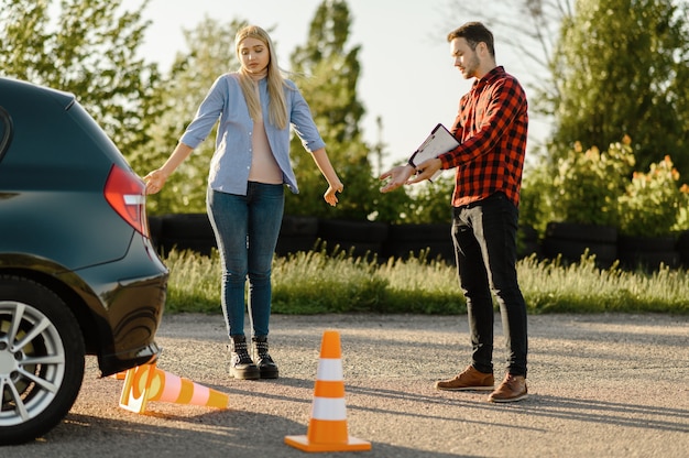 Male instructor and woman at the car and downed cone, lesson in driving school. Man teaching lady to drive vehicle. Driver's license education