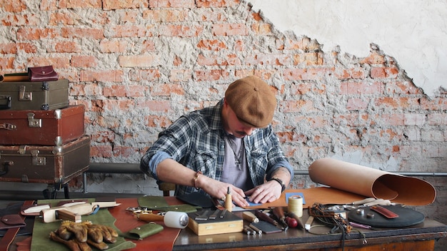 Male leather craftsman sews a leather wallet in the workshop. Manufacture of handmade leather products.