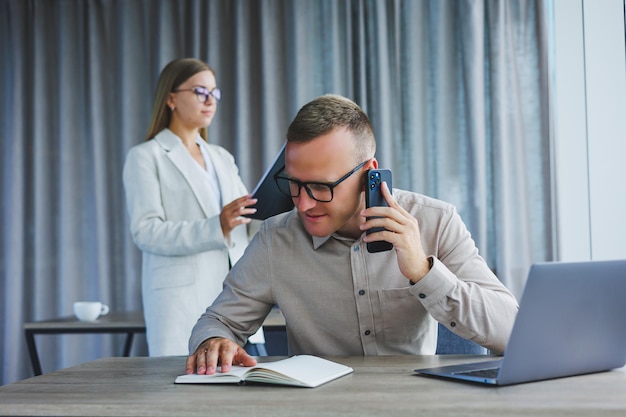 A male manager is talking on a mobile phone while sitting at a table with a computer and a notepad Working atmosphere in an office with large windows