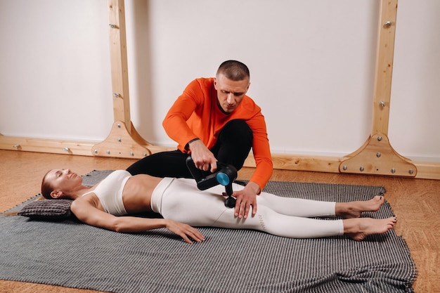 A male masseur massages a girl's body with a percussion massager to prepare for a stretching simulator