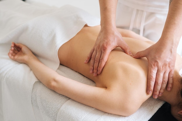 Male masseur massaging back and shoulder blades of young woman lying on massage table