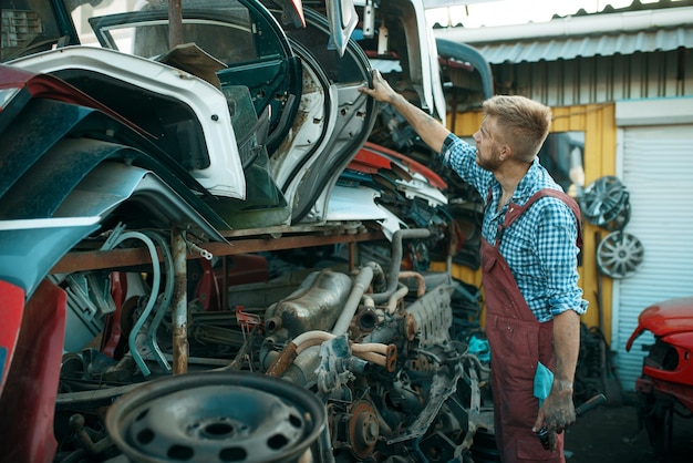 Male mechanic choosing door on car junkyard. Auto scrap, vehicle junk, automobile garbage. Abandoned, damaged and crushed transport, scrapyard