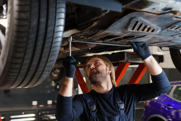 A male mechanic with a key unscrews the nuts on the bottom of the car that is on the lift Professional car maintenance in the service