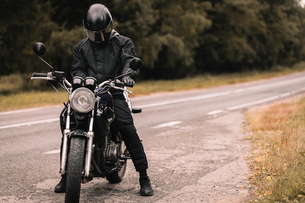 male motorcyclist in protective uniform and helmet with custom motorcycle cafe racer in autumn on th