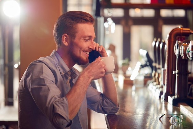 Male Owner Or Manager Of Bar Sitting At Counter Drinking Coffee And Talking On Mobile Phone